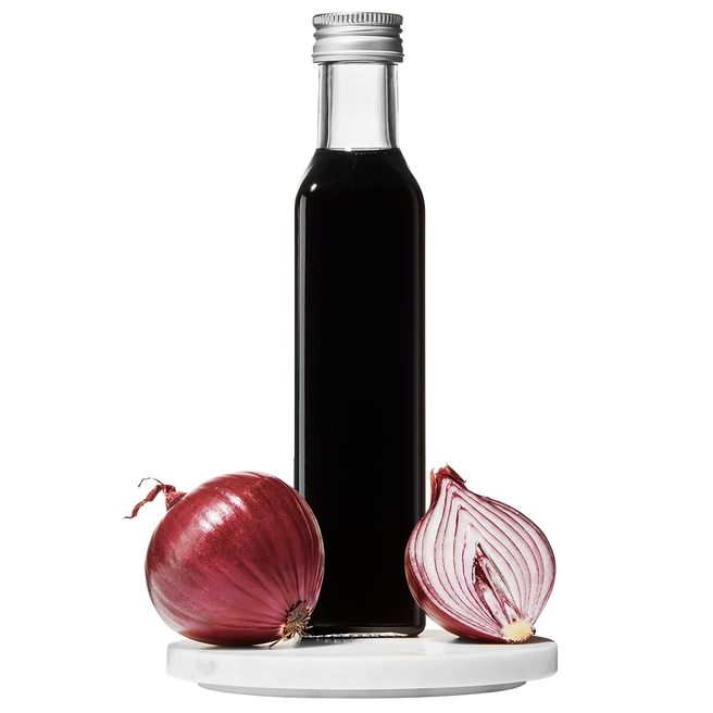 A clear glass bottle of Red Onion Balsamic Vinegar with a cap on top. A whole red onion and a slice of red onion are placed beside the bottle.