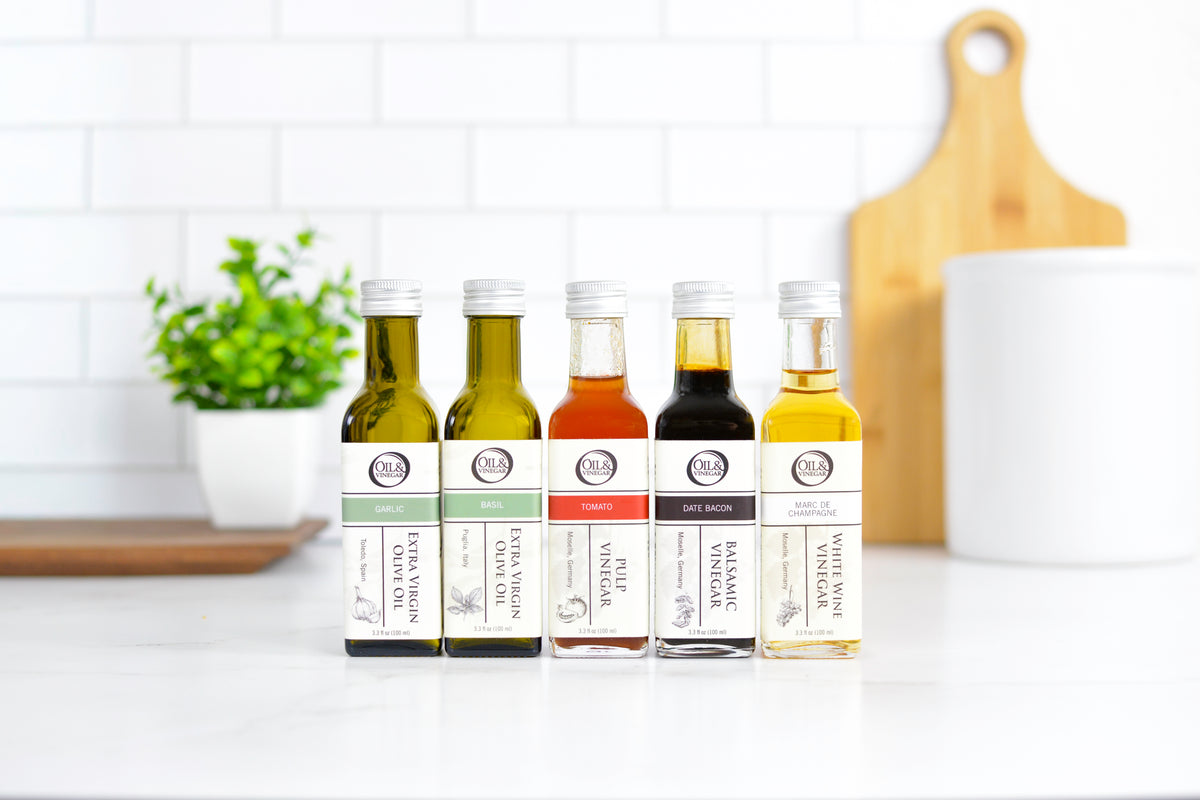 A gift set of five small bottles of spices or seasonings arranged in a gray box with crinkle paper filler on a white marble-like surface. The bottles have white labels with black text and are photographed from above with kitchen props around them.