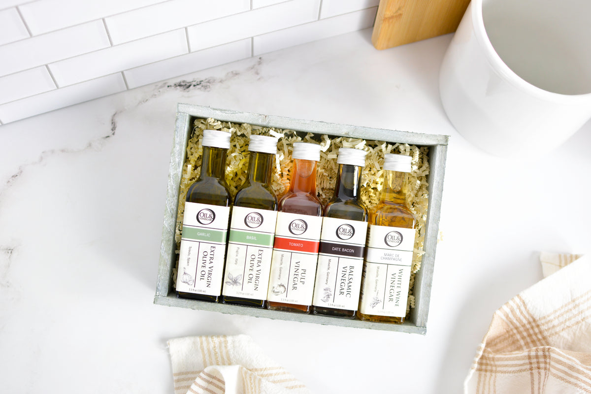 A gift set of five small bottles of spices or seasonings arranged in a gray box with crinkle paper filler on a white marble-like surface. The bottles have white labels with black text and are photographed from above with kitchen props around them.