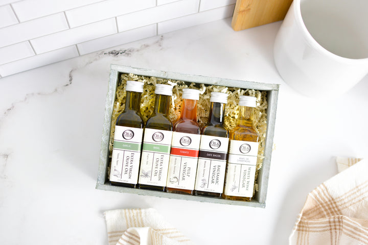 A gift set of five small bottles of spices or seasonings arranged in a gray box with crinkle paper filler on a white marble-like surface. The bottles have white labels with black text and are photographed from above with kitchen props around them.