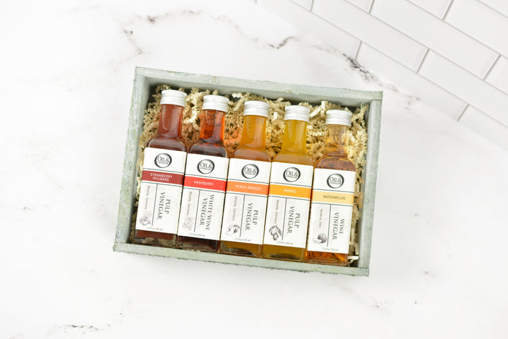 A gift set of five 100ml vinegar bottles in a wood tray with shredded paper filler, displayed on a white marble surface with a white subway tile background.
