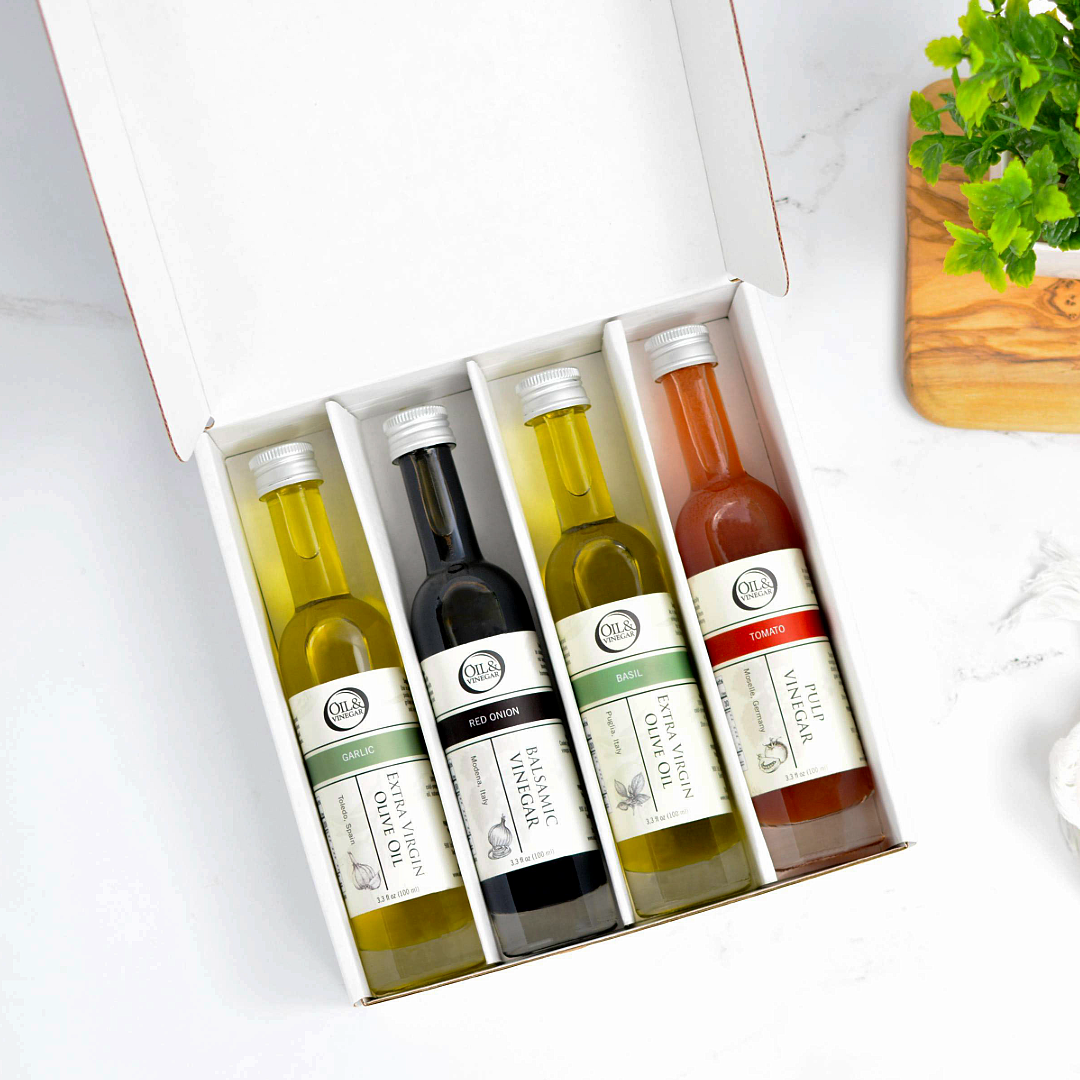 An open white gift box containing four small bottles of olive oil and vinegar. The bottles are arranged side by side inside the box, photographed on a light marble-like surface with a small plant and wooden board partially visible at the edges.