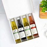 An open white gift box containing four small bottles of olive oil and vinegar. The bottles are arranged side by side inside the box, photographed on a light marble-like surface with a small plant and wooden board partially visible at the edges.