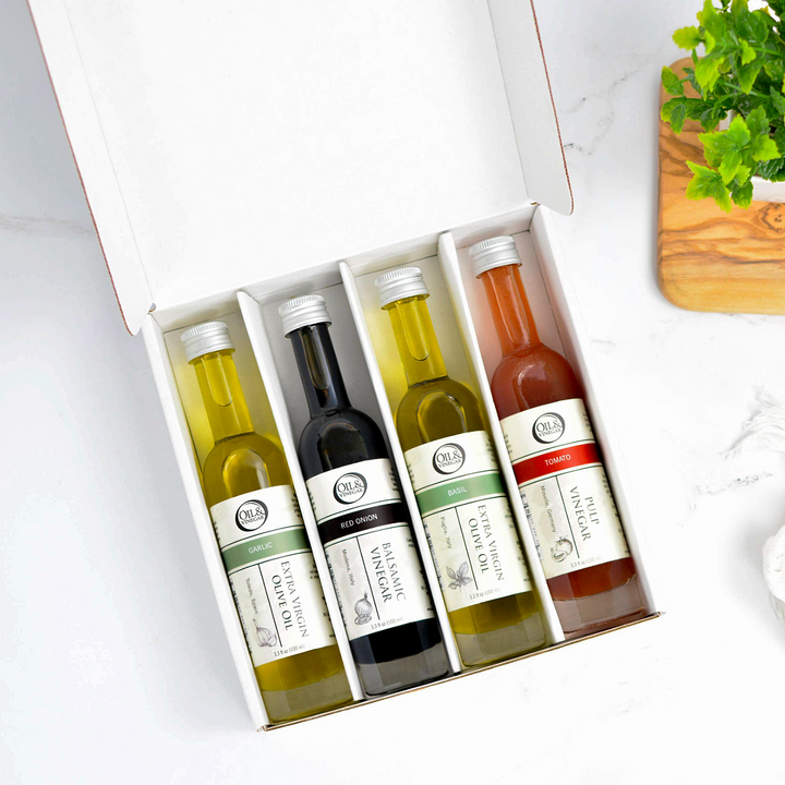 An open white gift box containing four small bottles of olive oil and vinegar. The bottles are arranged side by side inside the box, photographed on a light marble-like surface with a small plant and wooden board partially visible at the edges.