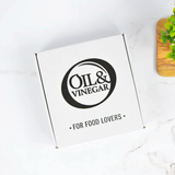 A white square box with a black circular logo reading 'Oil & Vinegar' and the phrase 'For Food Lovers' on top, photographed on a light marble-like surface with a small green plant in a wooden holder at the edge.