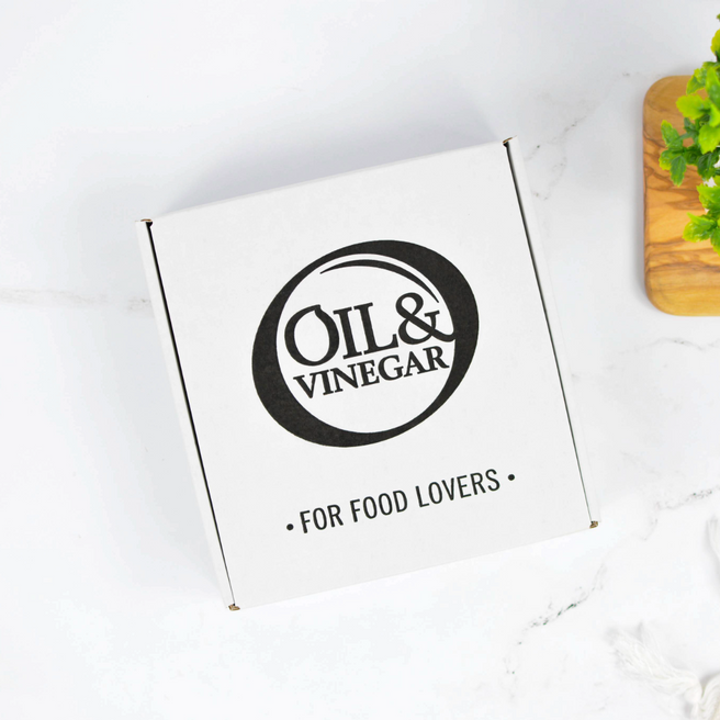 An open white gift box containing four small bottles of olive oil and vinegar. The bottles are arranged side by side inside the box, photographed on a light marble-like surface with a small plant and wooden board partially visible at the edges.