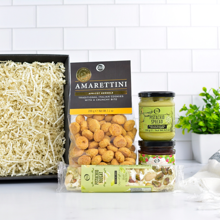 A gourmet gift box of Italian snacks and spreads, featuring a bag of Amarettini crackers, pistachio spread, and more arranged in a black tray with shredded paper filler.