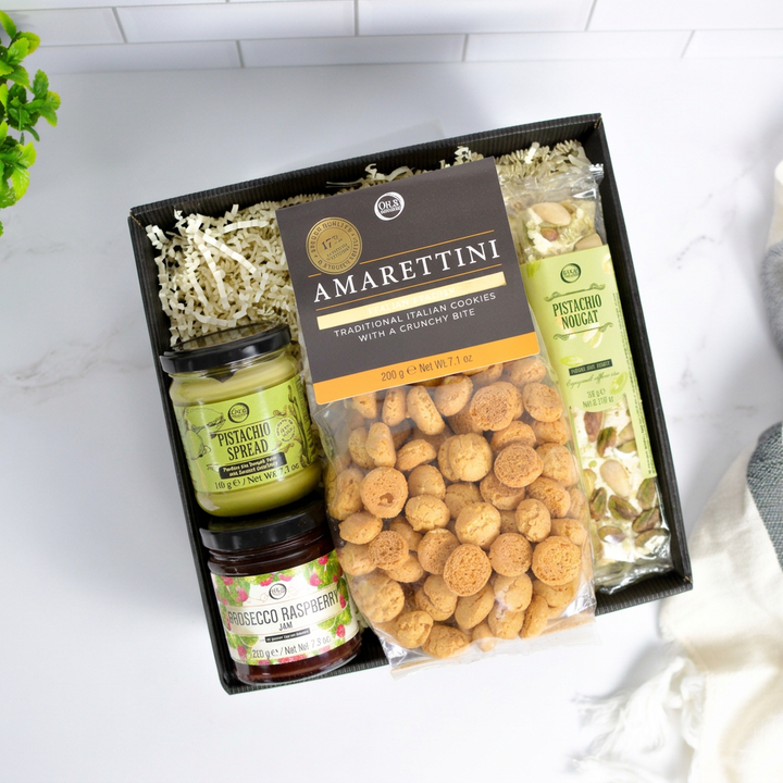 A gourmet gift box of Italian snacks and spreads, featuring a bag of Amarettini crackers, pistachio spread, and more arranged in a black tray with shredded paper filler.