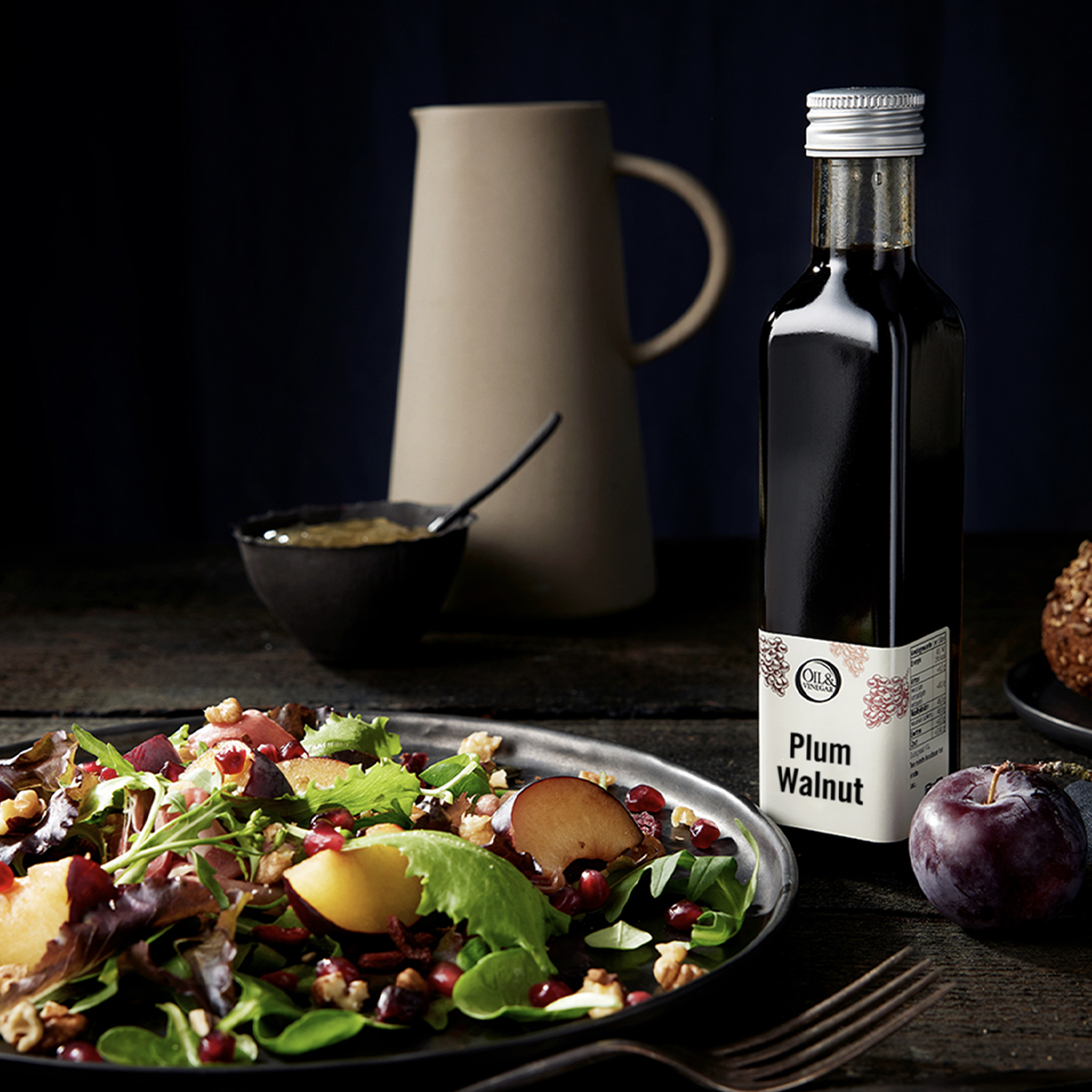 Plum Walnut vinegar bottle next to a salad plate with plums, pomegranates, and lettuce on a dark background
