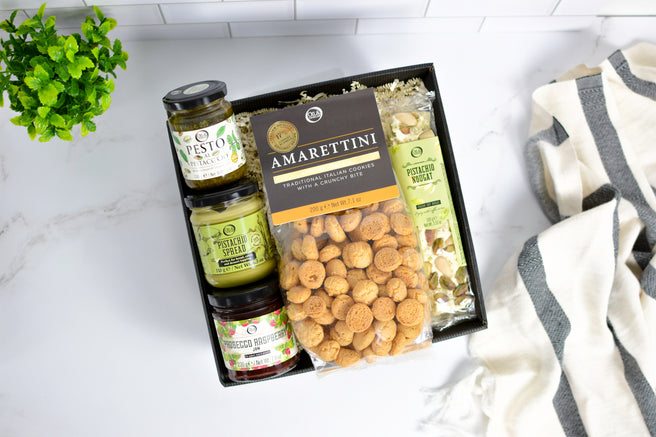 Gift box with Amaretti cookies, nougat, pistachio spread, pesto, and jam on a white surface with a plant and towel.