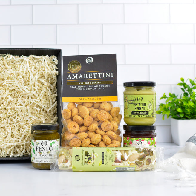 Gift box with Amaretti cookies, nougat, pistachio spread, pesto, and jam on a white surface with a plant and towel.