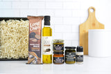 A curated food gift set arranged on a white countertop against a white subway tile backsplash. The set includes Long Chips Truffle chips, a bottle of olive oil, three jars of spreads, and a small bottle of truffle powder, and a wooden cutting board in the background.
