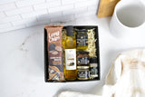 A gourmet gift box set on a white marble countertop, featuring a bag of Long Chips Nacho Potato Snacks, a bottle of Truffle Oil, and several small jars of gourmet spreads nestled in shredded paper inside a black box.