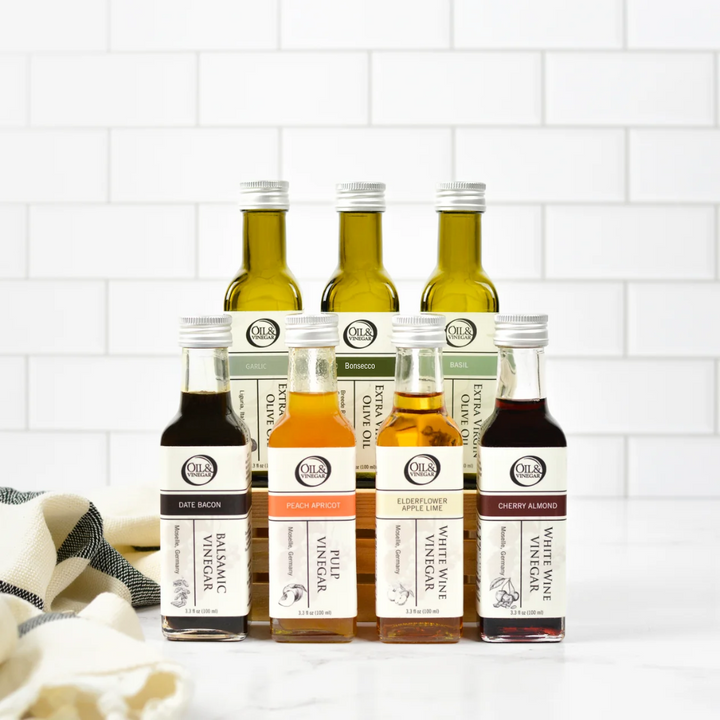 Seven small olive oil and vinegar bottles are lined up on a white countertop against a white subway tile backsplash. The bottles include various flavors and types of olive oil and vinegars, each with its own label and silver cap.