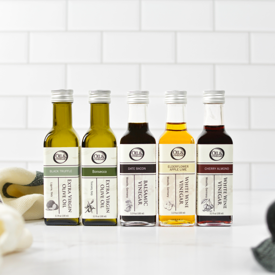 Five small olive oil and vinegar bottles are lined up on a white countertop against a white subway tile backsplash. The bottles include various flavors and types of olive oil and vinegars, each with its own label and silver cap.