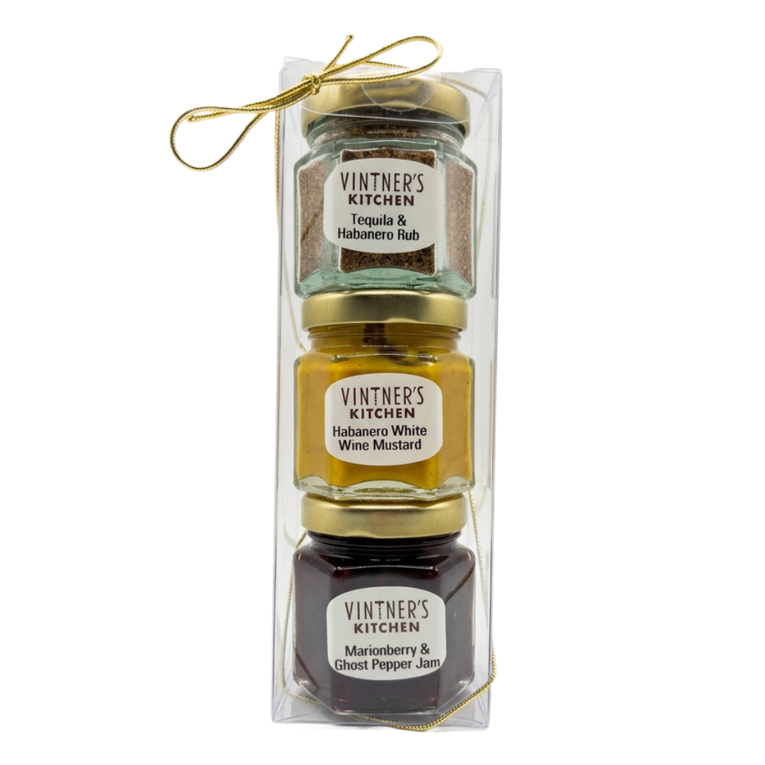 A clear plastic gift box containing three small glass jars of Vintner's Kitchen spreads, tied with a thin gold string. The top jar is Tequila & Habanero Rub, the middle is Habanero White Wine Mustard, and the bottom is Marinated Berry & Ghost Pepper Jam.