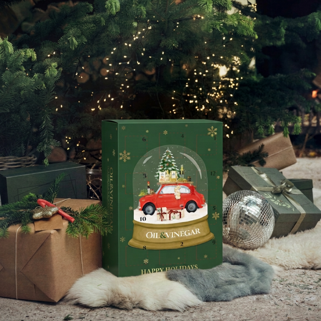A Christmas advent calendar box with a dark green background, gold snowflakes, and a central illustration of a red car inside a snow globe with a Christmas tree and gifts. The box is labeled 'OIL & VINEGAR' and 'HAPPY HOLIDAYS'.