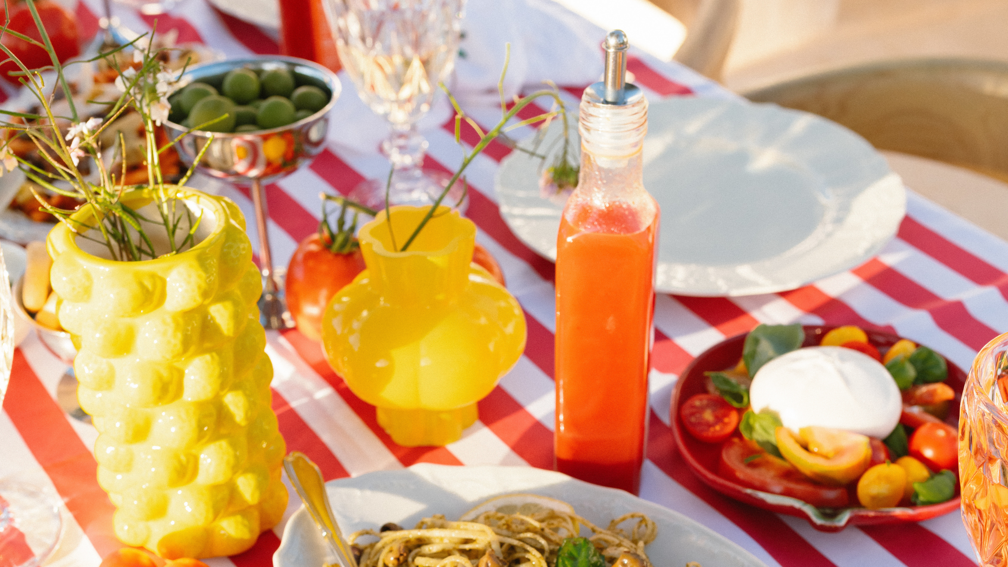 Outdoor dining table with pasta, salad, and drinks on a red and white checkered tablecloth.